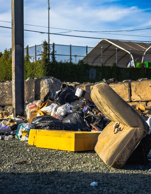 A pile of mixed household rubbish and waste items is scattered on the ground next to a stone wall in an outdoor setting. The waste includes several black plastic garbage bags, a yellow plastic contain