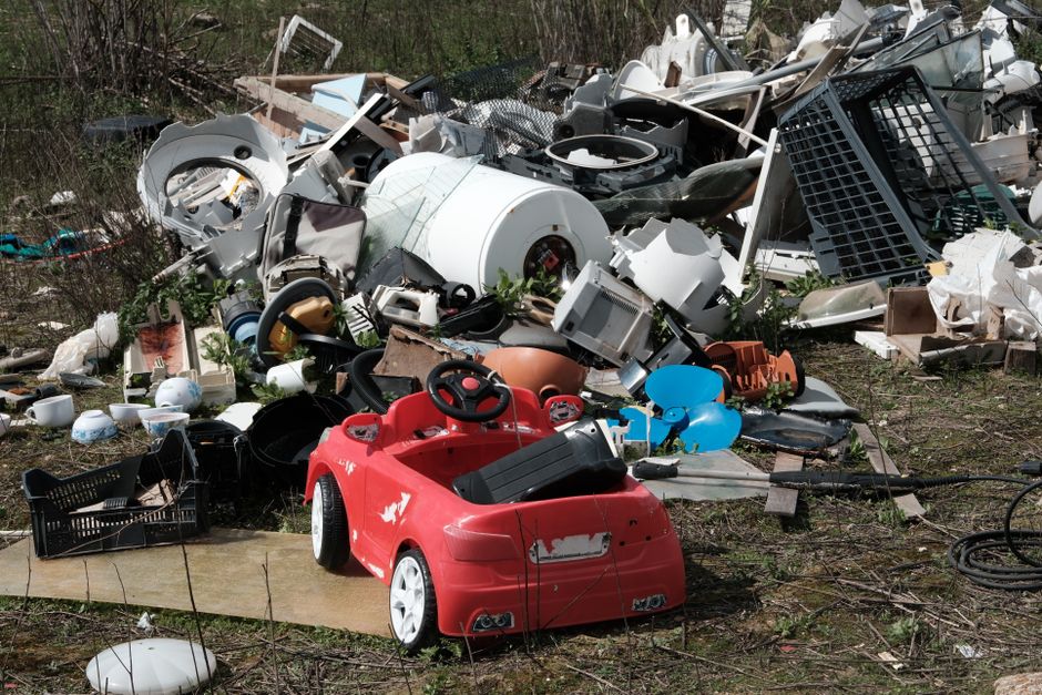 A large excavation site with steep, sloped banks composed of exposed soil and layers of mixed waste materials, including discarded plastic bags, fabric pieces, and other refuse, some of which are blue